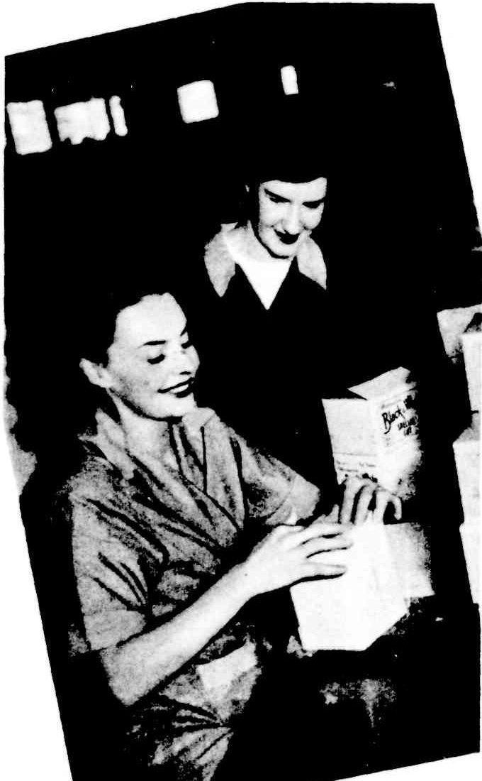 A young woman seated at a bench sealing a cigarette box while her supervisor looks on.