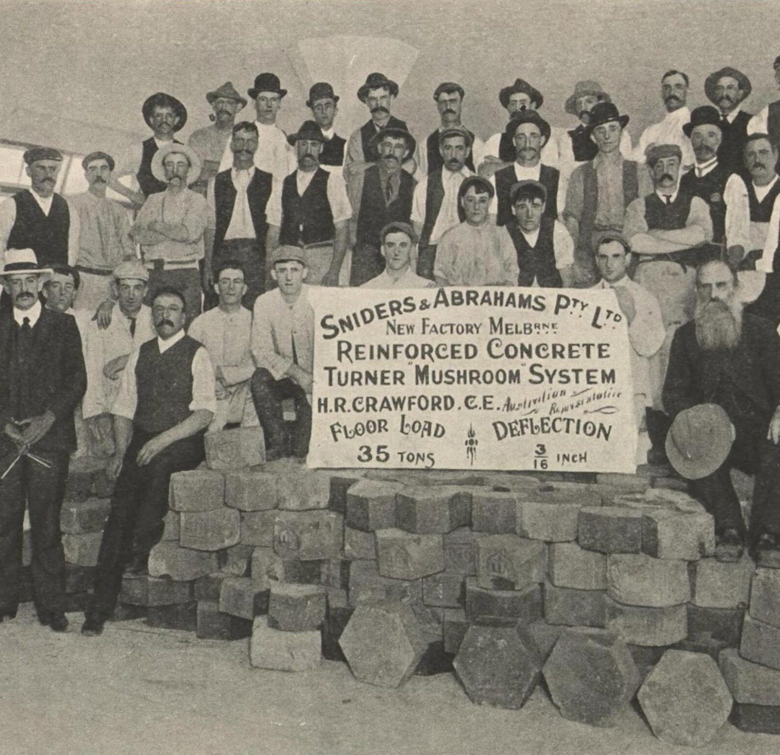 About forty men posed on and around a heap of stacked blocks on the new factory floor, behind a sign recording a 35-ton load.