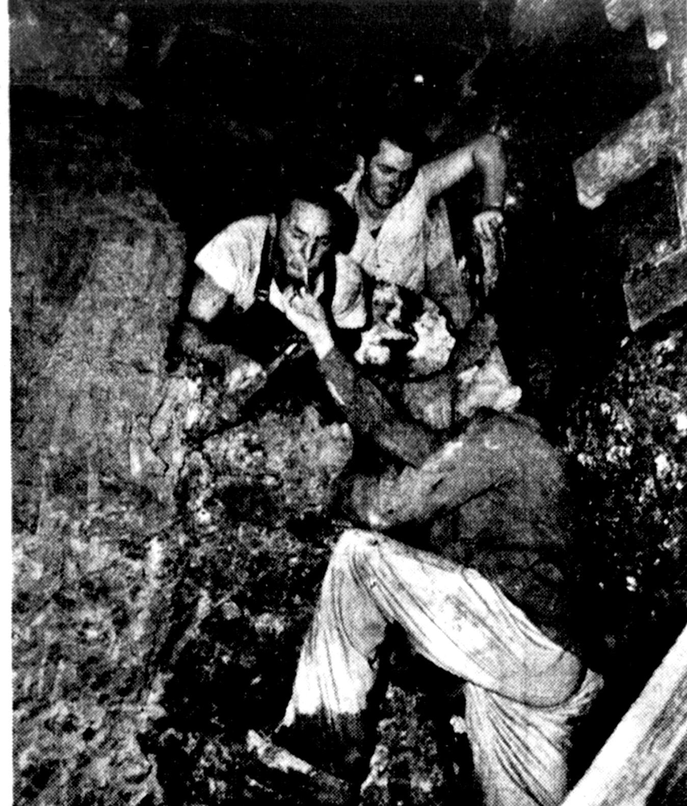 Tunnellers crouched in a dark tunnel of earth and timber, two of them sharing a light for a cigarette while a third looks on.