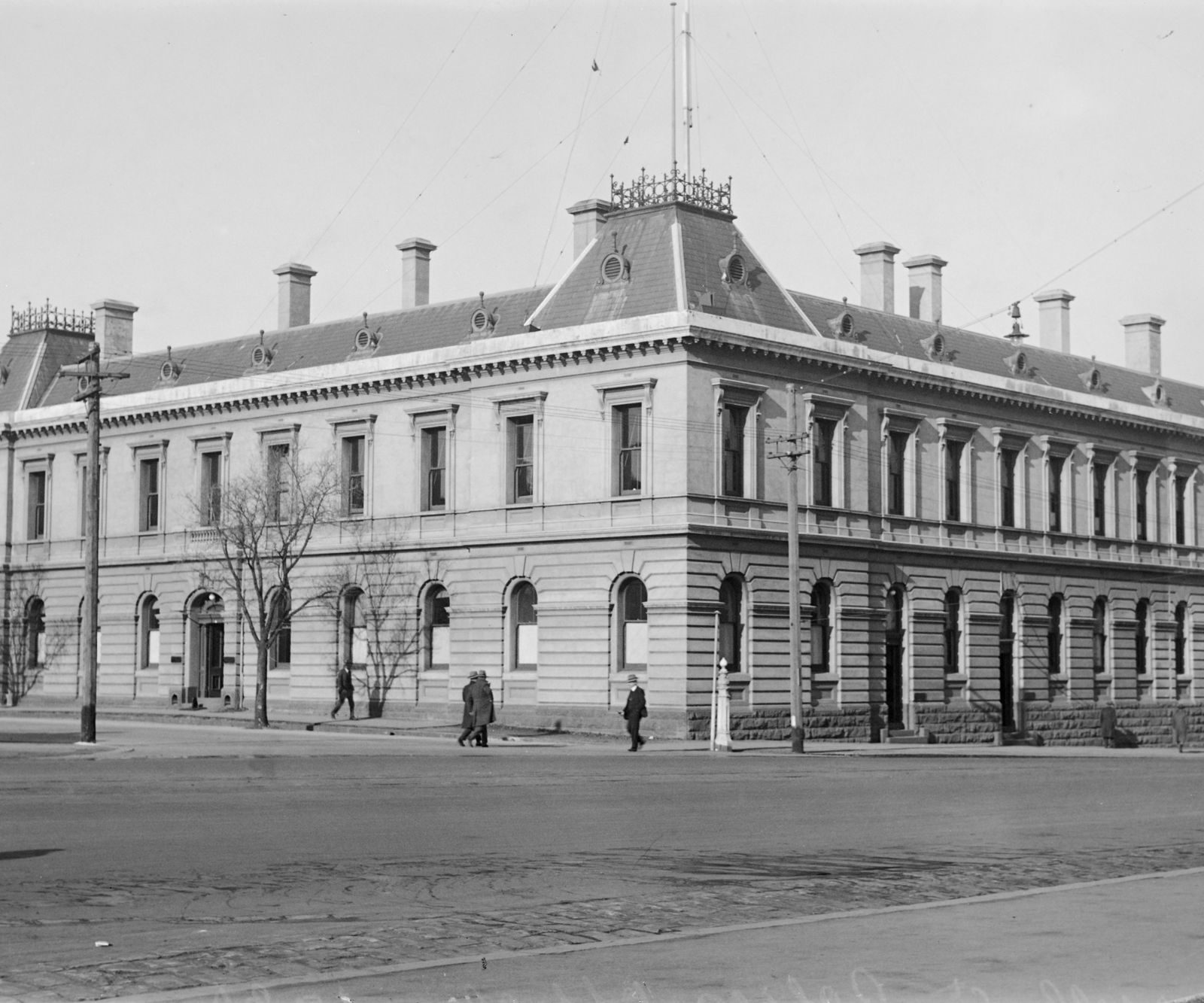 The two-storey police offices on a street corner around 1930, men in hats walking past.